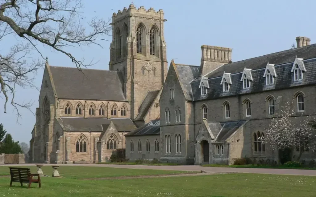 A large, historic stone building with arched windows and a central clock tower stands surrounded by green lawns and trees under a clear sky. Benches sit on the grass in the foreground.